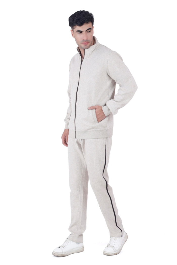 A man dressed in Turfit’s Oatmeal Millage tracksuit with black vertical piping, standing in a casual pose with one hand in the pocket and head turned slightly, wearing white sneakers against a white background.